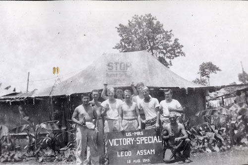 Soldiers of the 748th Railway Operating Battalion pose outside of tent holding a Ledo road sign while celebrating Victory over Japan Day in the Pacific during World War II.  Part of the research collection of the U.S. Army Transportation Museum.
