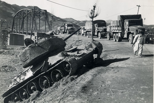 Forward Transportation in Korea.  US Army trucks pass a Korean using an A-Frame to porter materials to the front.  Both a passing a knocked out North Korean T-34 tank.