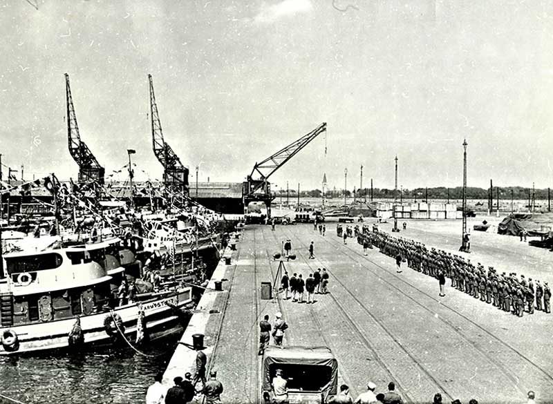 An Army company standing in formation on a shipping dock lined with tug boats.